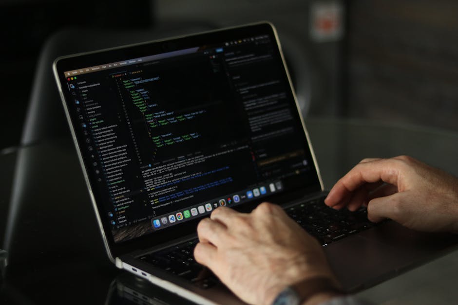 A person operates a laptop computer placed on a dark desk, with visible code and text on the screen, indicating a workspace focused on digital or technical tasks. The individual's hands are positioned